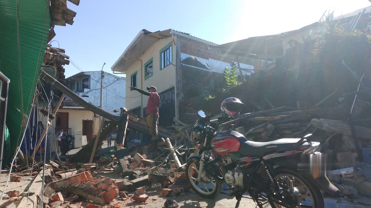 Eine Person auf umgestürzten Balken zeigt auf ein beschädigtes zweistöckiges Haus; eingestürzte Ziegelmauern und Schutt umgeben ein geparktes Motorrad im Vordergrund.