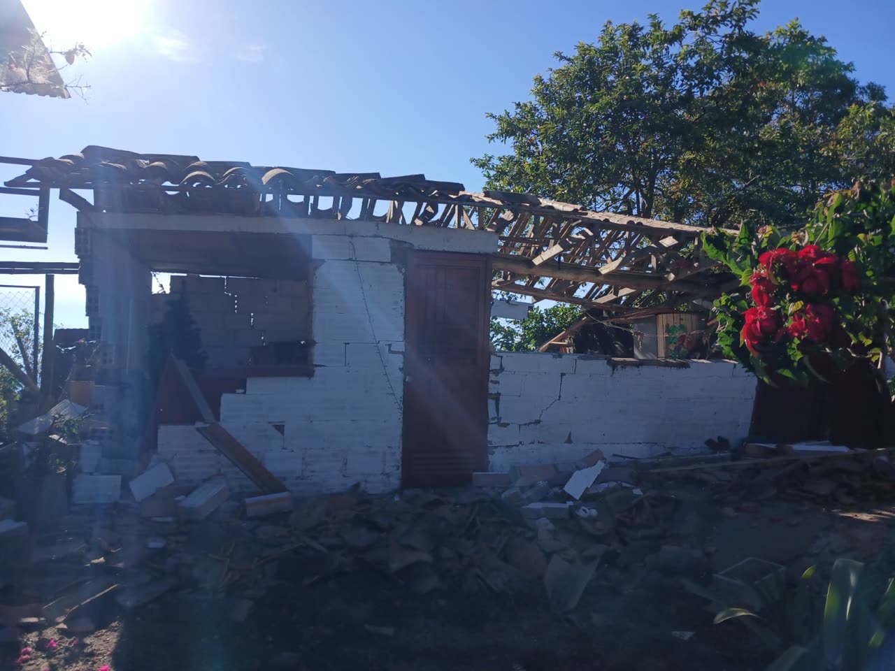 Ein einstöckiges Haus mit zerstörten weissen Blockwänden und eingestürztem Ziegeldach, das Holzgerüst gegen den Himmel freigelegt; Trümmer bedecken den Hof, rechts wachsen rote Bougainvillea-Blüten.