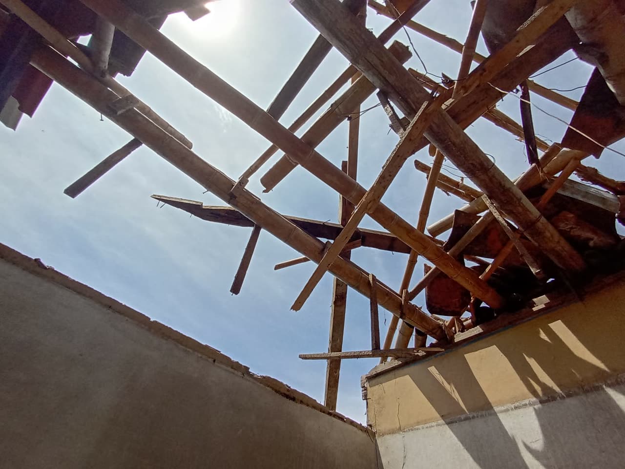 Blick aus einem Zimmer nach oben auf ein zerstörtes Dach: gekreuzte, zersplitterte Bambusstangen und Bretter hängen über den Wänden, dahinter offener Himmel.