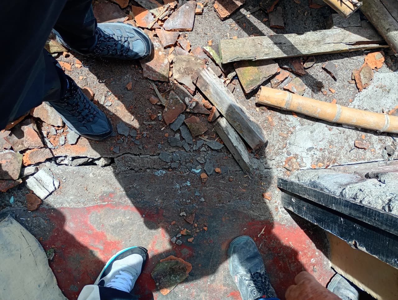 Von oben gesehen die Schuhe mehrerer Personen, die zwischen zerbrochenen Dachziegeln, Brettern und einer Bambusstange auf einem rissigen Boden stehen.