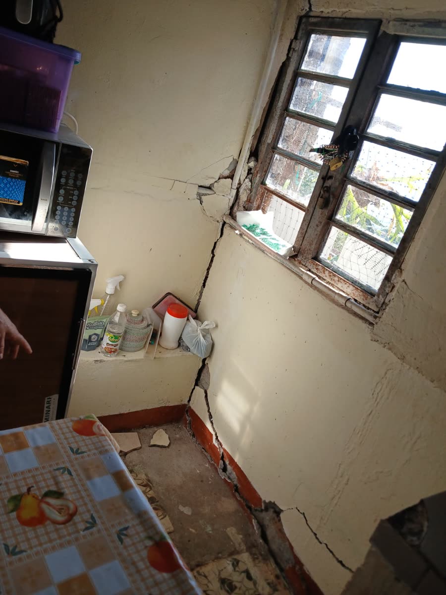 Küchenecke mit einem breiten diagonalen Riss, der vom Fensterrahmen die verputzte Wand hinunterläuft; in der Nähe eine Mikrowelle, Putzflaschen und eine gemusterte Tischdecke.