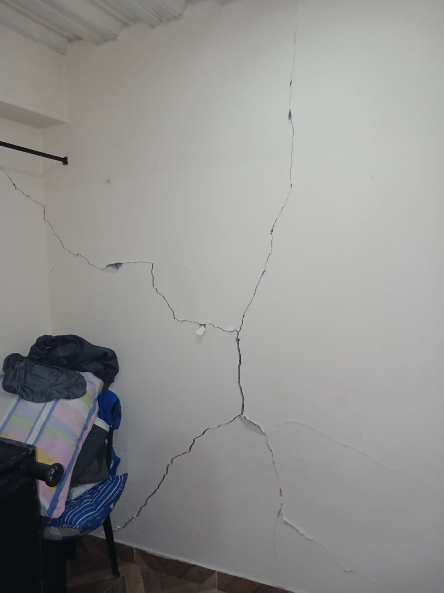 Weisse Schlafzimmerwand mit einem langen, sich verzweigenden Riss von der Decke bis zum Boden; in der Ecke gefaltete Kleidung und ein Stuhl.