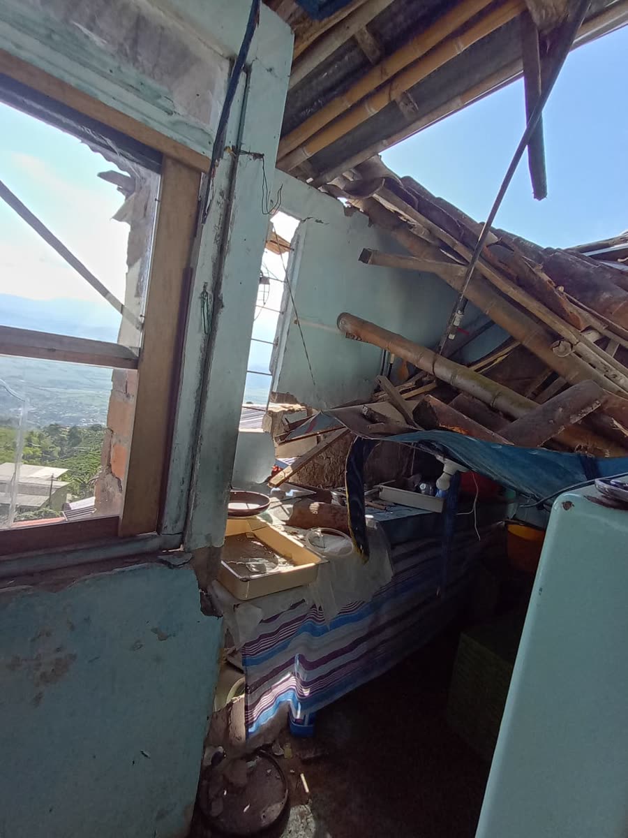 Eine Küche mit nach innen eingestürztem Dach: Bambusstangen und Bretter liegen über der Arbeitsfläche, der Fensterrahmen ist gebrochen, und wo die Wand fehlt, sind grüne Hügel zu sehen.