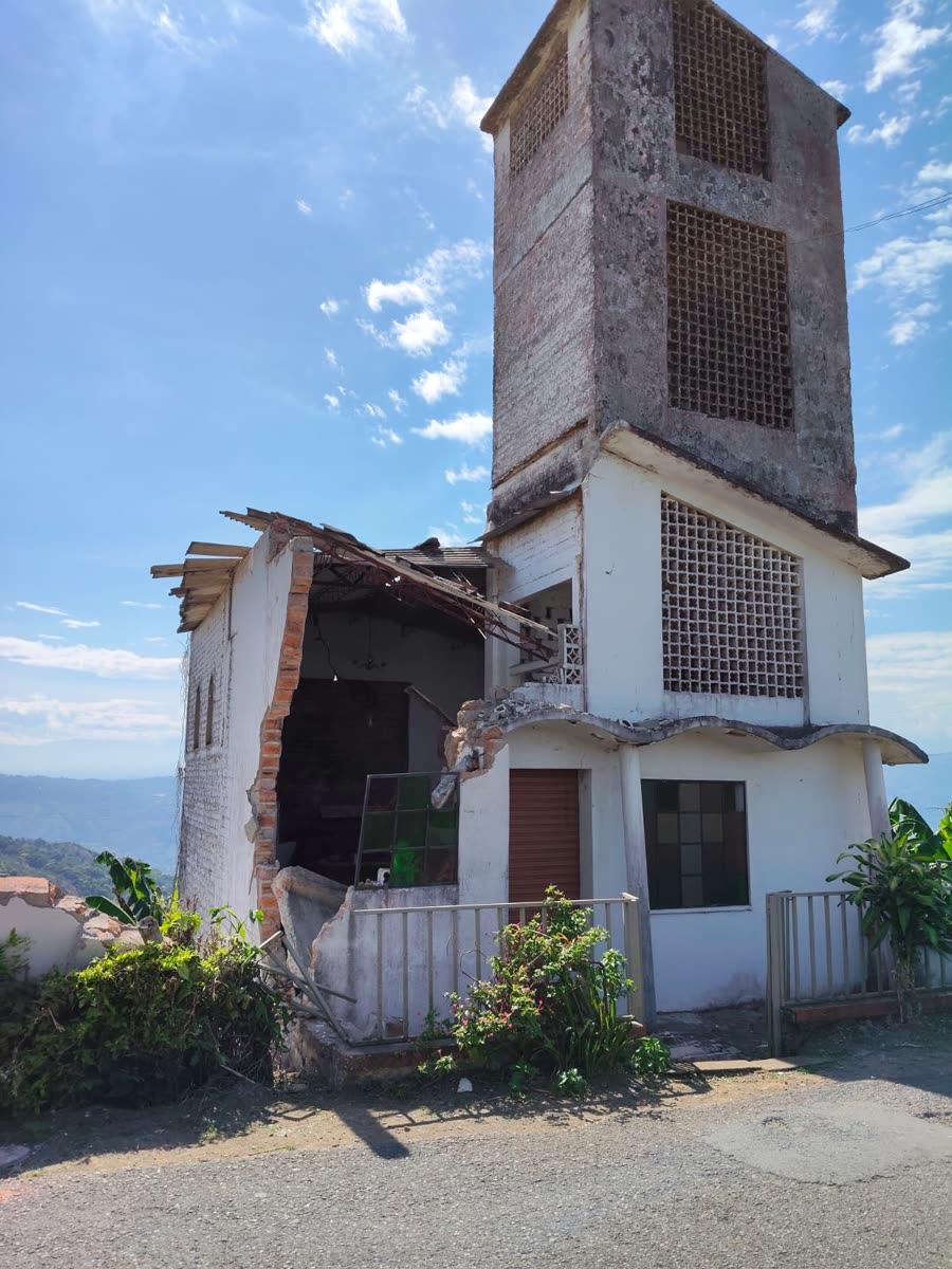 Ein weisses zweistöckiges Haus mit hohem Gitterziegel-Turm; die gesamte Seitenwand ist weggebrochen und gibt ein Zimmer mit Hängelampe frei, dahinter ein grünes Bergtal.
