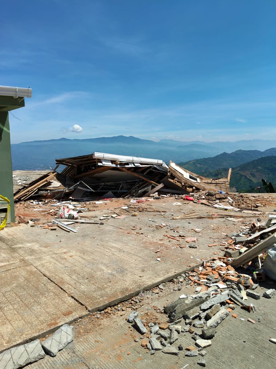Die flach liegenden Überreste eines Hauses — Dachbleche, Holz und verstreute Ziegel — auf einer Betonterrasse, in der Ferne dunstige blaue Bergketten.
