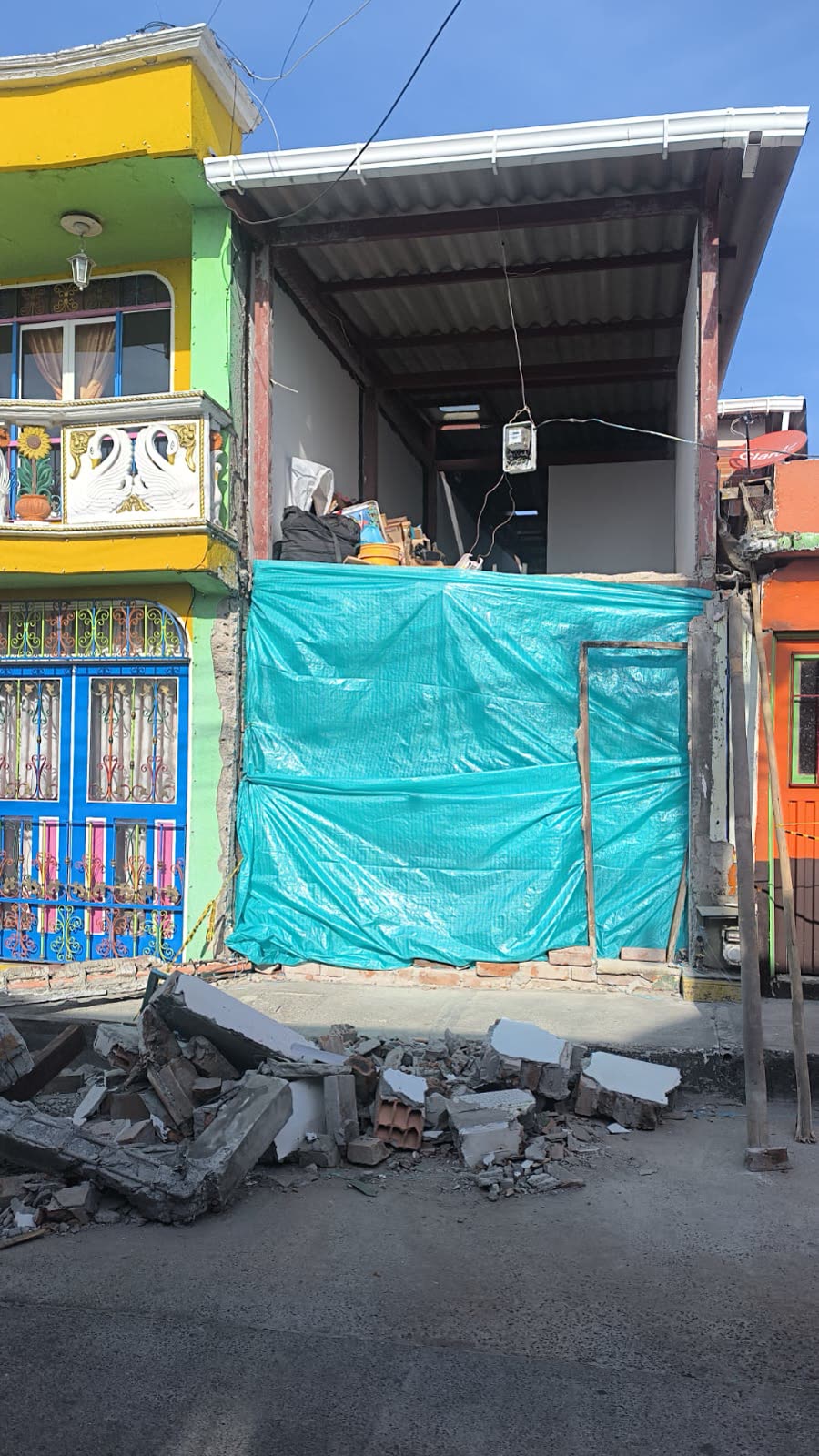 Eine Lücke zwischen einem gelben Haus mit verziertem Schwanen-Balkon und einem orangen Haus: Das Haus dazwischen ist nur noch ein Rohbau, mit türkiser Plane verschlossen, davor aufgehäufter Schutt.
